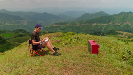 Hombre-Sentado-Al-Aire-Libre-En-La-Cima-De-Una-Colina-Viendo-La-Televisión-Antigua-Y-Tomando-Notas