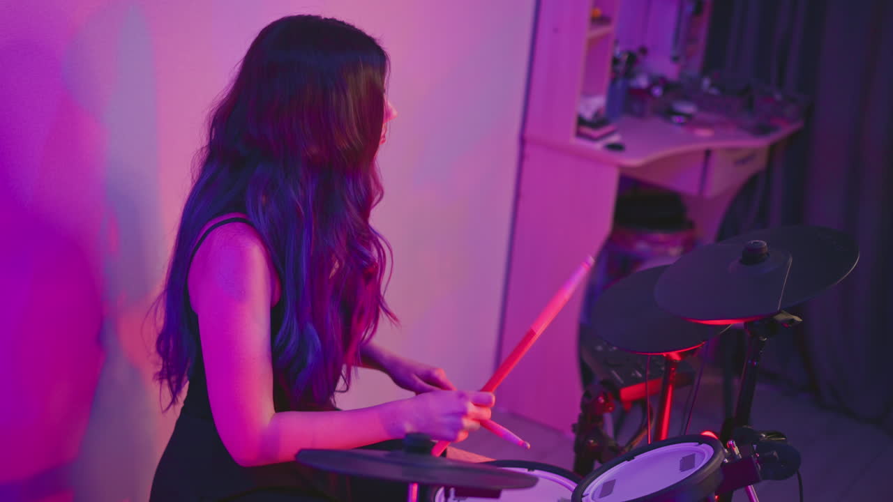 Woman with long hair casually practicing on electronic drum set in softly lit studio, holding drumsticks while seated near wall, background features shelf and curtain with gentle purple-pink glow