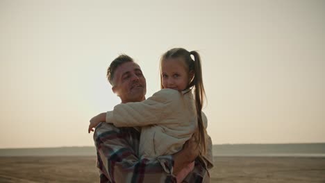 Zoom-in:-Portrait-of-a-little-blonde-girl-in-a-white-jacket-who-is-held-in-her-arms-by-her-father,-a-middle-aged-brunette-man-with-gray-hair-in-a-checkered-shirt-during-his-vacation-outside-the-city-in-the-summer-evening