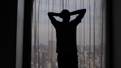 Silueta-De-Un-Hombre-Estirándose-Y-Mirando-Por-La-Ventana-Con-Las-Cortinas-Cerradas,-Oscureciendo-La-Vista-Panorámica-De-La-Metrópolis-De-La-Ciudad