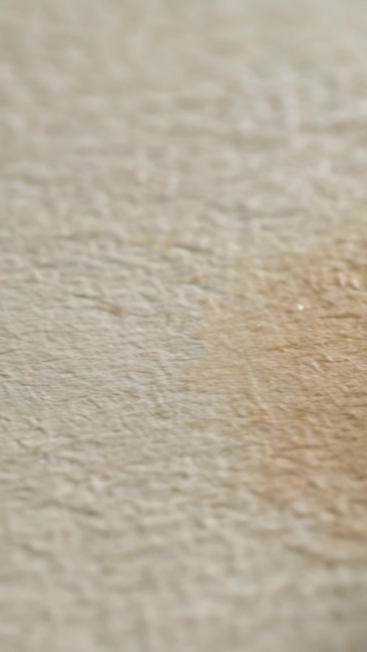 Video verticale: Una punta acuminata che entra, proiettando un'ombra verso una macchia marrone su tessuto beige, primo piano.