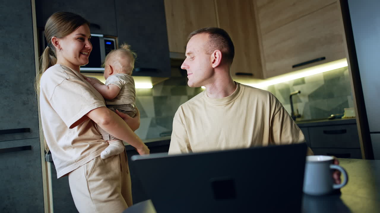 Long-haired woman with baby in hands stands near her husband. Man sitting at table works on laptop.