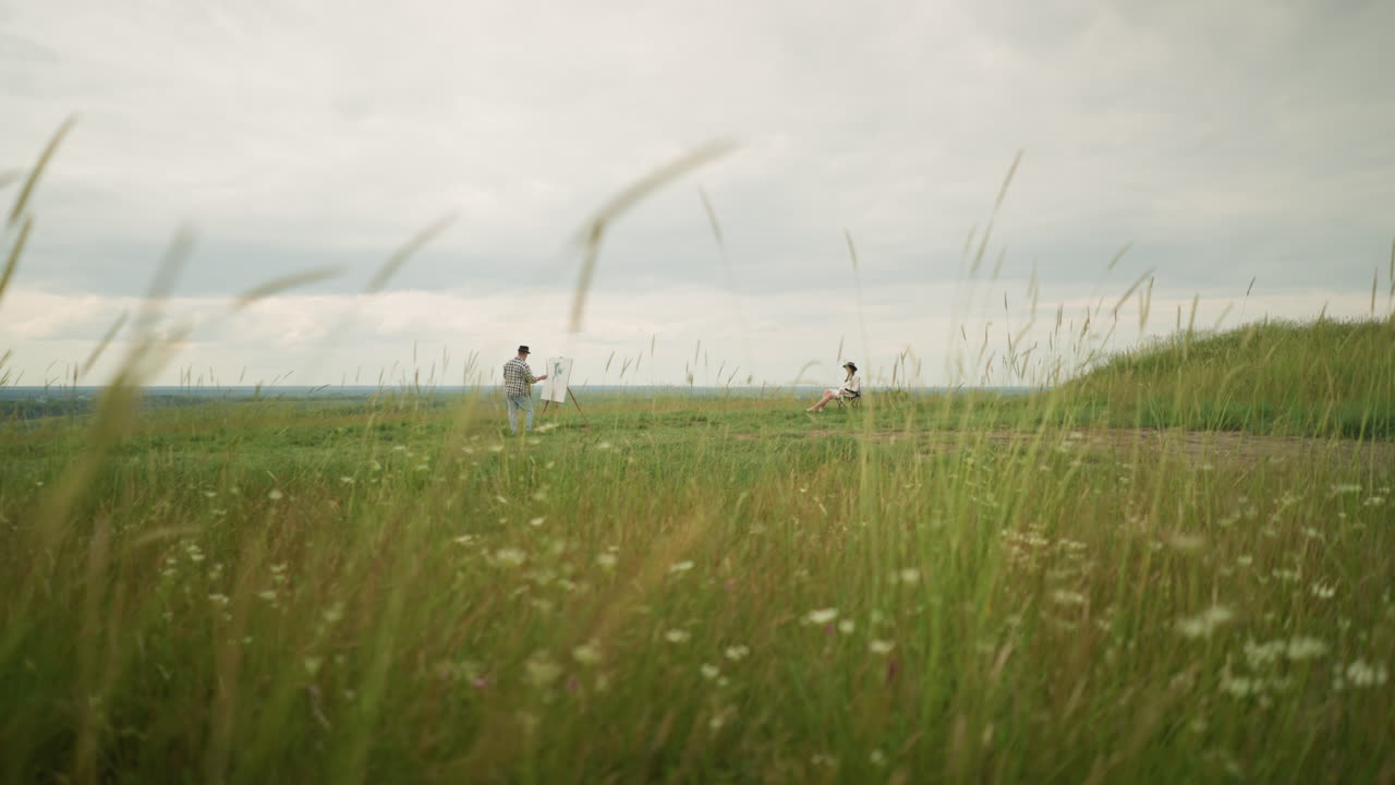 모자와 체크색 셔츠를 입은 예술가는 구름이 은 하늘 아래 높은 잔디의 조용한 에서 기둥 위에 그림을 그리고, 한 여인이 인근 의자에서 휴식을 취하는 동안, 자연의 포옹 속의 평화로운 창의성