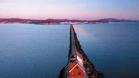 aerial footage flying towards shore past the brick lighthouse at the end of a snow dusted rocky breakwater in rockland maine at sunrise