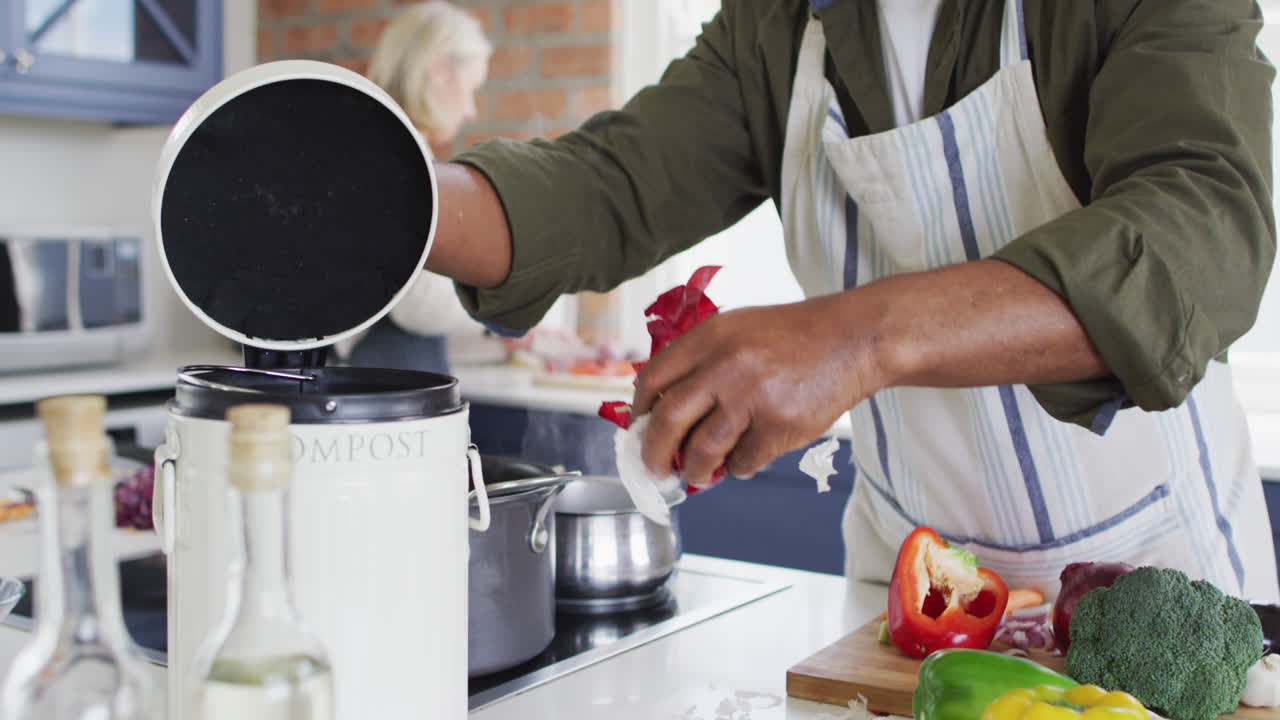 hombre mayor afroamericano con delantal poniendo cáscaras de verduras en un contenedor de compost en la cocina