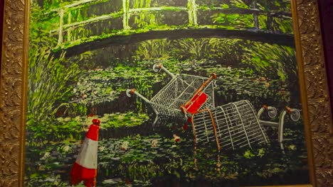 an oil painting made by banksy at his exhibition in tallinn estonia showing shopping carts or trollies and traffic cones thrown in a river that has a bridge going over it. art in a golden frame.