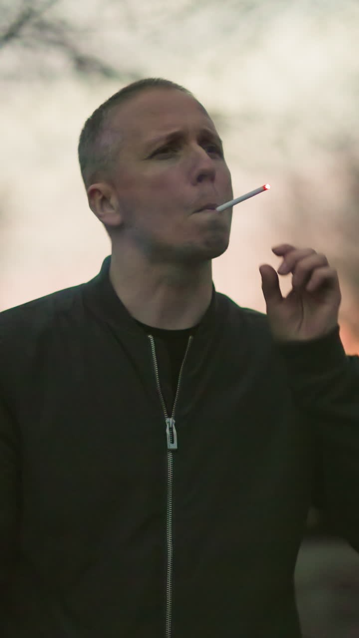 человек в зеленой куртке зажигает сигарету в сумерках в тускло освещенном лесу, на заднем плане изображена размытая машина с включенными фарами