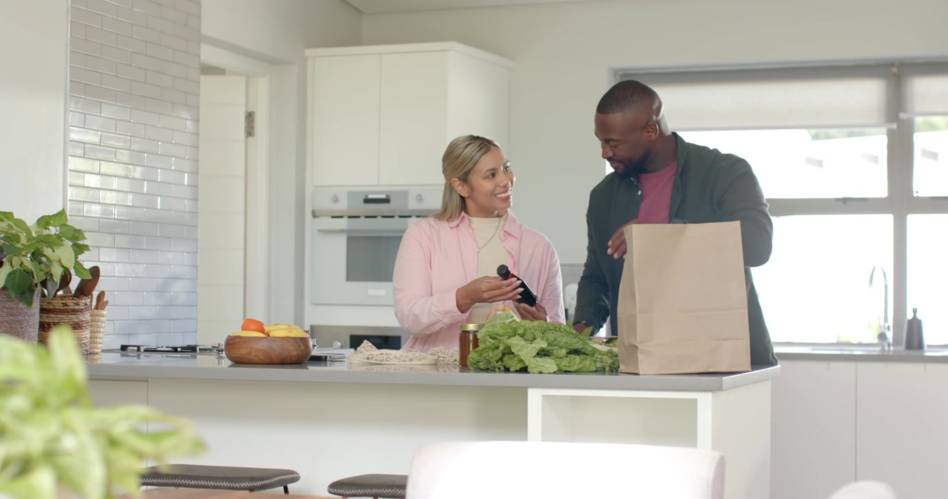 Pareja afroamericana desempacando comida en la cocina, el hombre llega con una bolsa de papel, leyendo la etiqueta.