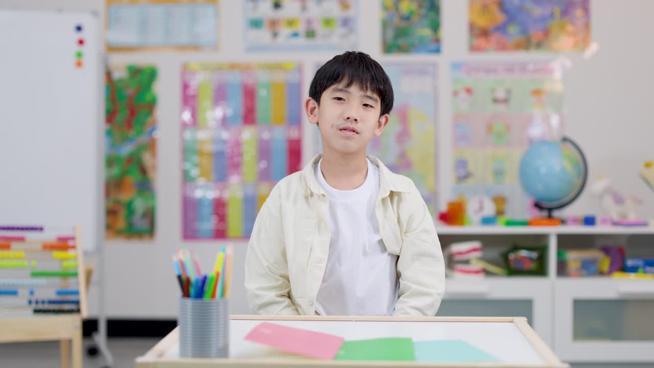 明るくカラフルな教室で腕を組み、自信ありげに机に向かうアジアの小学生の男の子