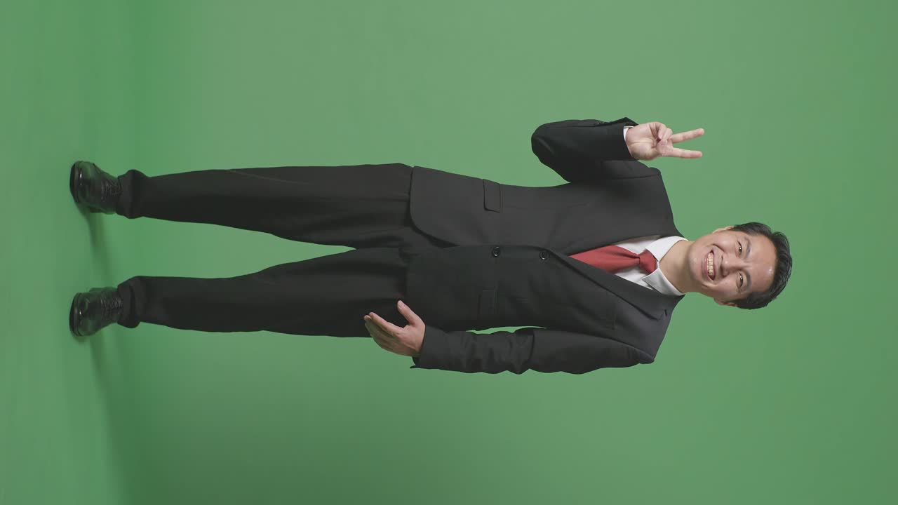 아시안 사업가의 몸매가 가득 차 있으며, 스튜디오의 녹색 스크린에 서서 평화와 미소를 짓고 있습니다.