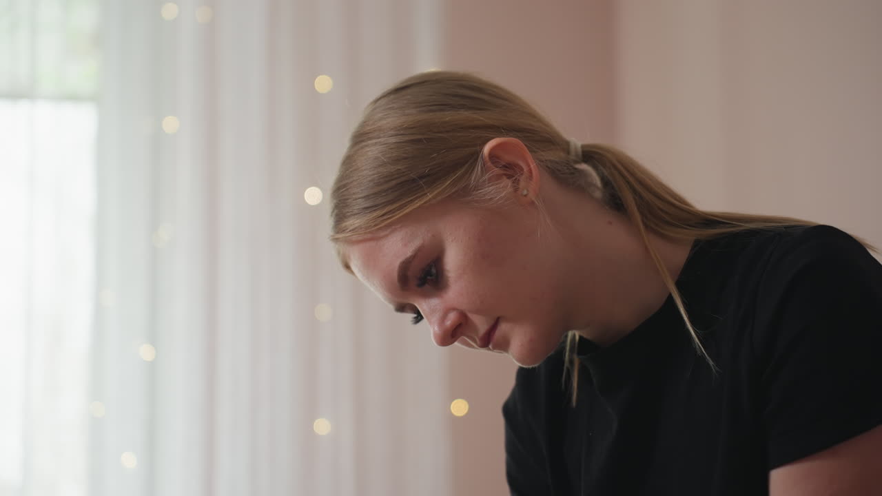Light skin blonde woman with hair parted at middle and tucked behind ear, wearing black shirt, looking down with calm expression, white curtain and soft daylight in background