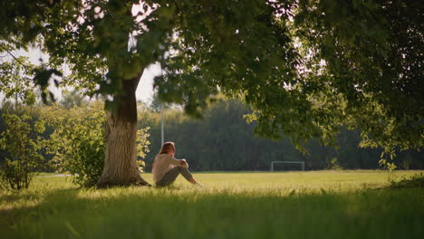 woman sits under tree with arms folded over her legs, gazing thoughtfully into distance, gentle sunlight filters through swaying leaves,with background greenery and distant goalpost on open field