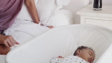 millennial mother sitting on bed looking at her newborn baby sleeping in a crib,tilt shot