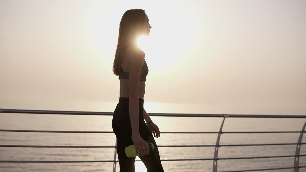 아름답고 우아한 소녀가 스포츠 옷을 입고 산책로를  ⁇ 고 있습니다. 손에 스포츠 병을 들고 있습니다.  ⁇ 습니다. 태양이 비추고 있습니다. 슬로우 모션