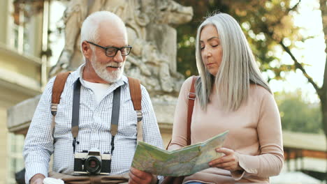 retrato de um casal de turistas seniores sentados no monumento no centro de uma cidade, conversando e olhando para o mapa