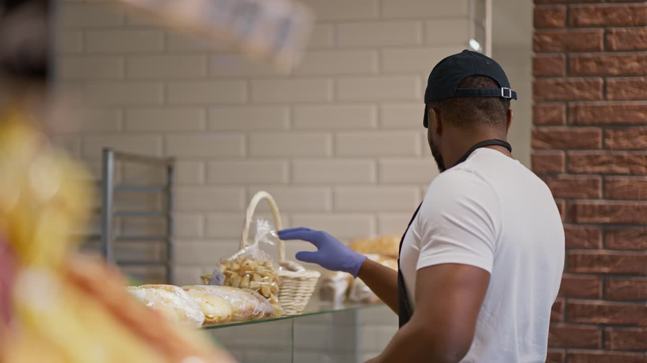 vista lateral de un hombre de piel negra trabajador feliz de un supermercado en una camiseta blanca y una gorra negra colocando pasteles y otros productos de panadería en una vitrina en un supermercato moderno