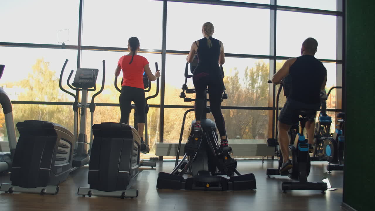 The view from the back: In the cardio area of the gym women on elliptical machines and a man on an exercise bike train near a large panoramic window in slow motion.