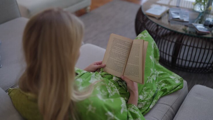 Vista da sopra la spalla di una donna che legge un libro in uno spazio interno tranquillo. La scena evidenzia concentrazione, relax e un momento di vita pacifico con illuminazione soffusa e un'atmosfera accogliente