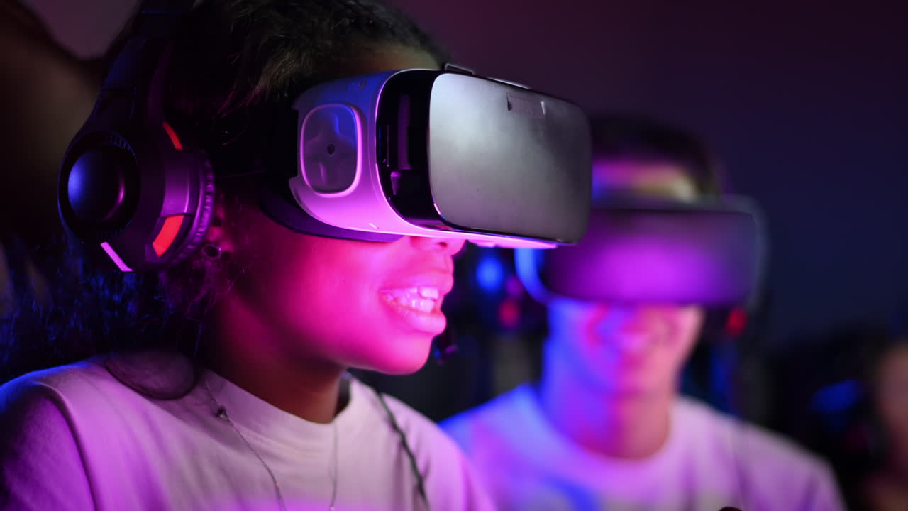 White boy and black girl teens in VR headsets playing video games in video game club with illumination using a gamepad, talking in voice chat. Slow motion