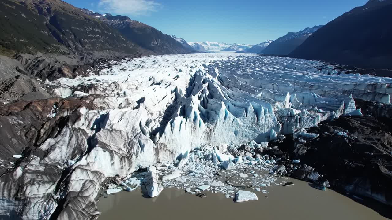 Aerial views showcase the stunning expanse of a glacier in Patagonia. The vivid blue ice contrasts with the surrounding mountains and waters under a clear summer sky, highlighting nature's beauty.