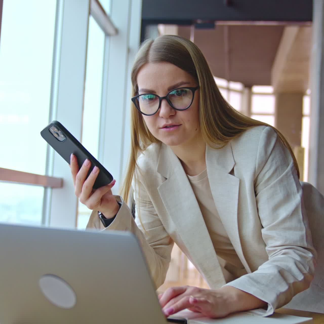 Long-haired woman in glasses uses phone and laptop at once. Lady stands above the desk and types something on computer