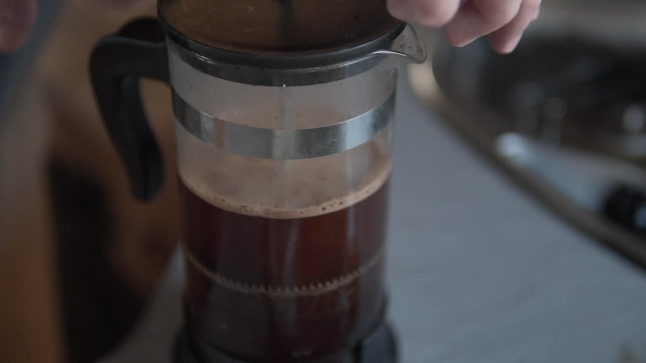 A close-up view of hands gently pressing the plunger of a clear glass French press, brewing rich, dark coffee with visible grounds in a cozy indoor setting