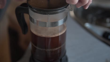 a close-up view of hands gently pressing the plunger of a clear glass french press, brewing rich, dark coffee with visible grounds in a cozy indoor setting