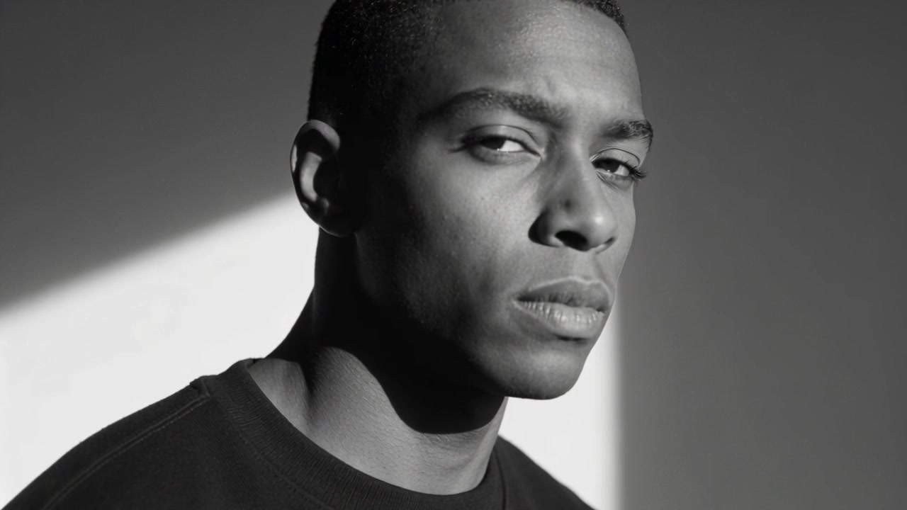 Mies kääntyy merkistä, katsoo kameraan studiossa, diagonaalisessa valaistuksessa, yllään tumma pyöreäkauluksinen paita.