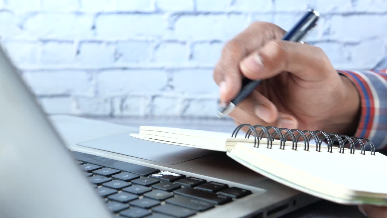 persona tomando notas en una computadora portátil con un bloc de notas