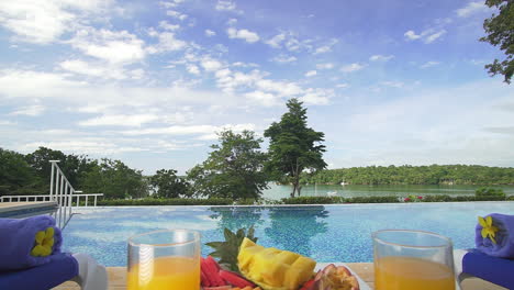 Hermosa-Vista-Junto-A-La-Piscina-Con-Dos-Vasos-De-Jugo-De-Naranja,-Vista-Sobre-El-Entorno-Natural-Con-Un-Ligero-Acercamiento