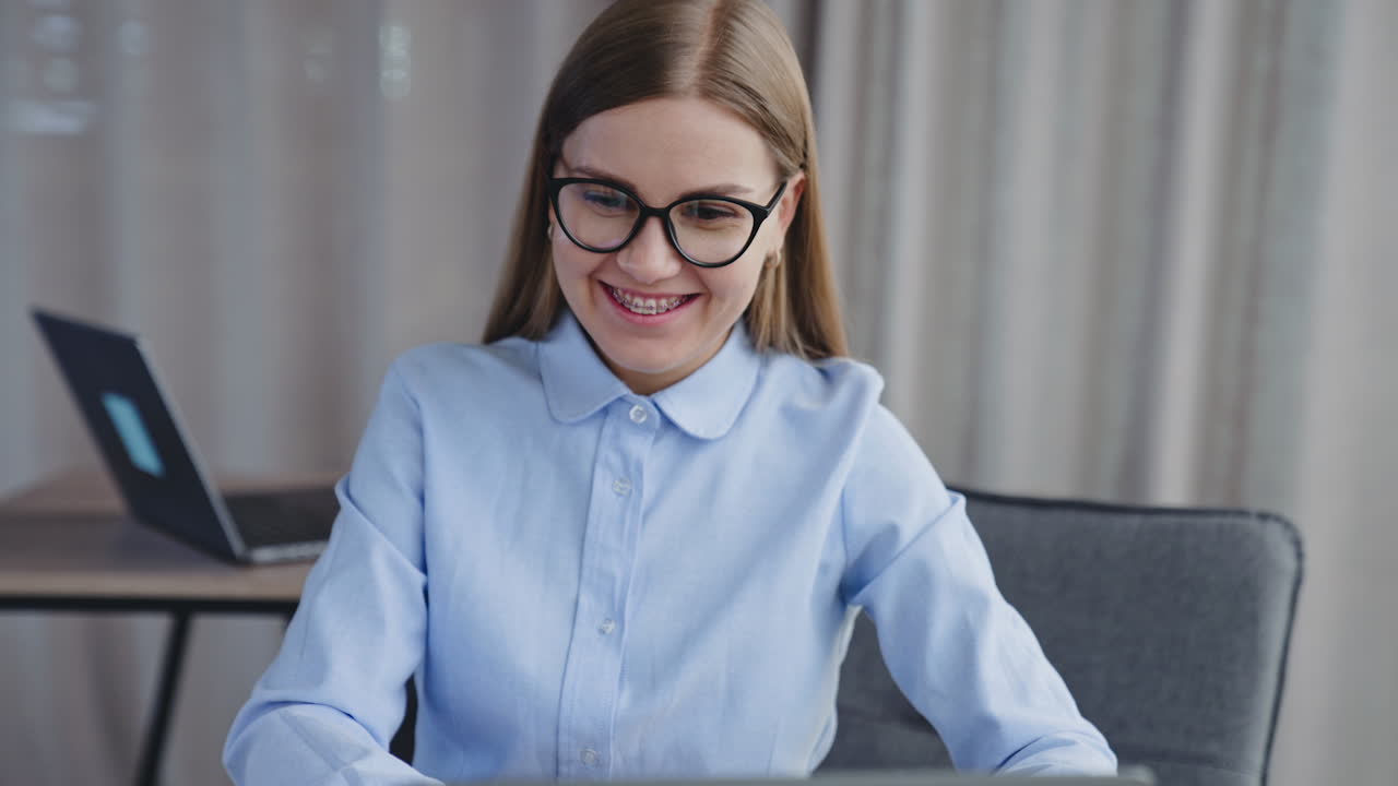 Happy smiling long-haired lady in glasses and braces on her teeth looks at the screen of computer. Cheerful employee in the office.