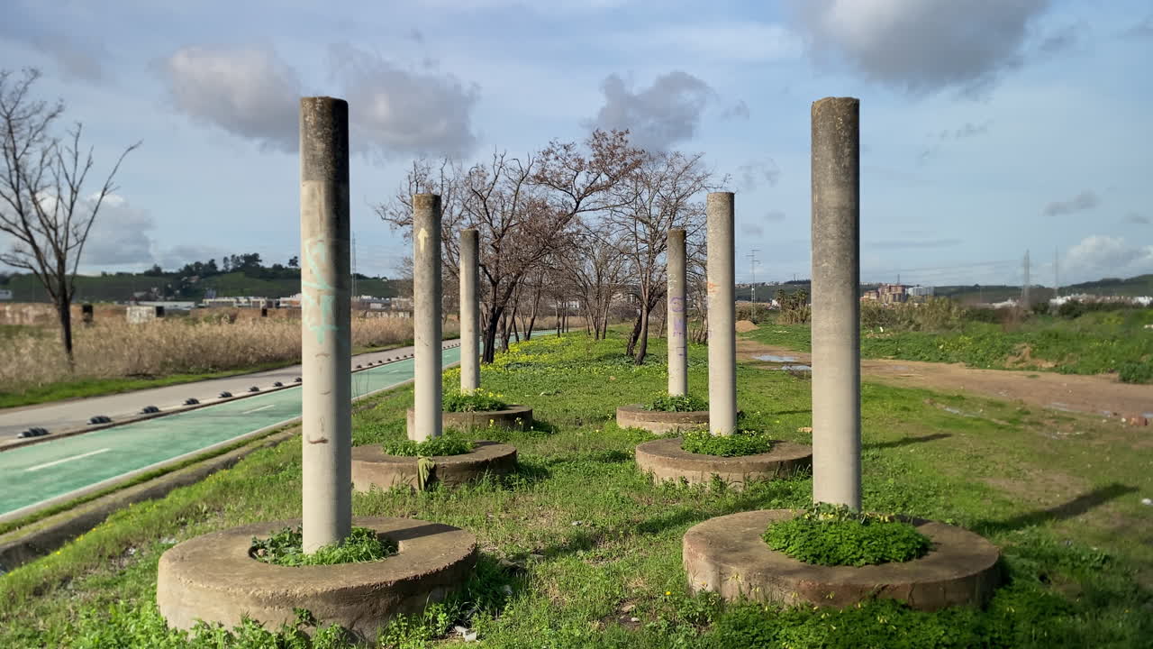 Pilastri di cemento che si ergono su un sentiero erboso lungo il fiume, simboleggiando il degrado urbano, l'edilizia e la resilienza contro gli elementi naturali