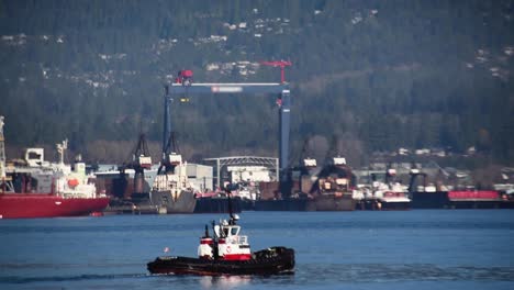 Canadian-mooring-boat-sailing-in-the-port-of-Vancouver-for-its-next-towing-with-a-high-blue-crane-of-a-shipyard-at-the-background