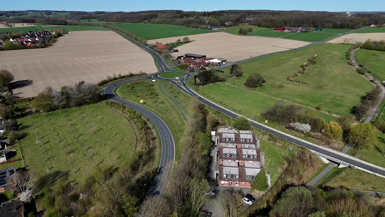Suburb residential apartments of town during sunny day in spring. Traffic on intersection road with roundabout. Farm fields on hill in german town. Aerial forward wide shot.