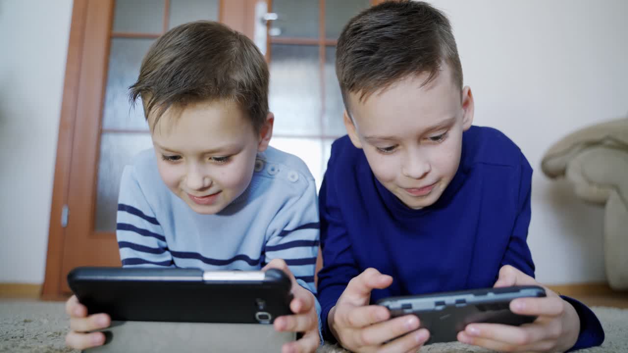 Happy boys playing on wireless gadgets while lying on the carpet at home. Elder boy helps his friend on a digital tablet. Close-up