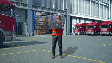 full body of asian male engineer with safety helmet working on a laptop and looking around while standing , outside of logistics distributions warehouse