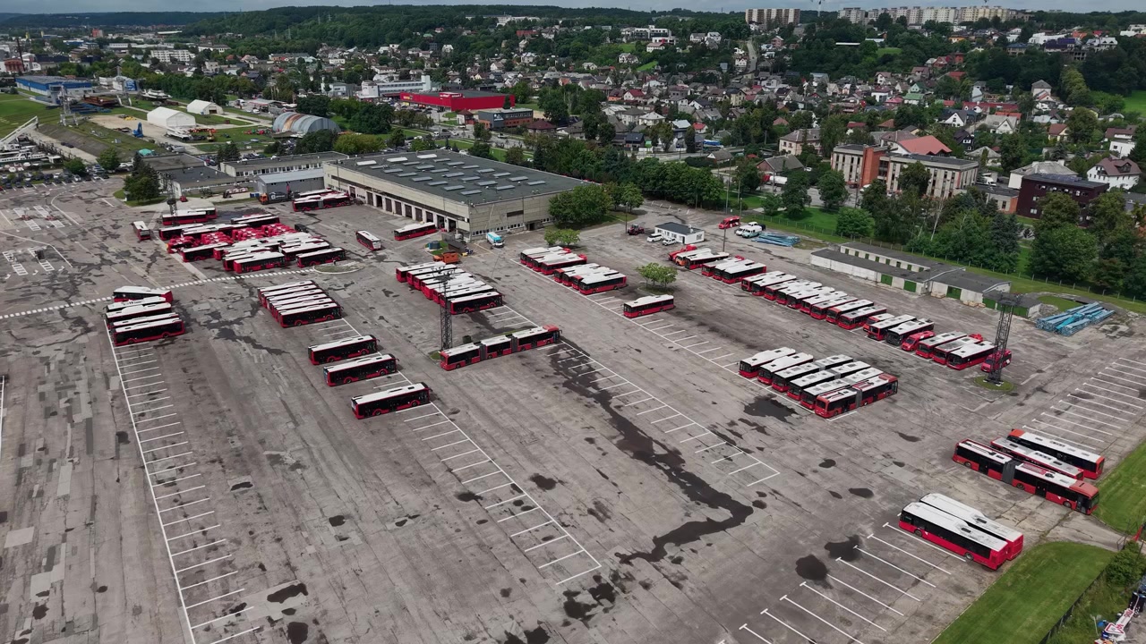 Rozległe ujęcia z lotu ptaka przedstawiają rozległy Park Autobusowy w Kownie z czerwonymi autobusami ustawionymi w równe rzędy, szerokimi betonowymi placami, miejskim otoczeniem, dramatycznymi chmurami i bez widocznych ludzi. Idealne dla transportu miejskiego.