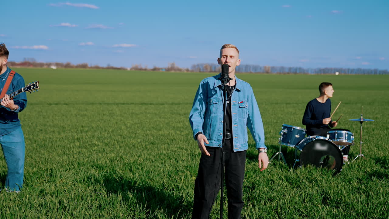 Blond guy singer at the microphone in nature. Music band playing musical instruments while man soloist singing on green field in sunny day.