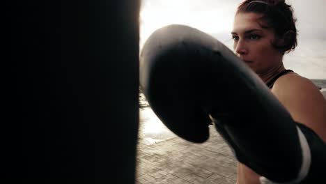 Vista-De-Cerca-De-Una-Boxeadora-Fuerte-Con-Guantes-Golpeando-Una-Bolsa-De-Pie-Contra-El-Sol-Junto-Al-Mar.-Boxeadora-Entrenando-Temprano-En-La-Mañana-En-La-Playa.-Filmado-En-4k