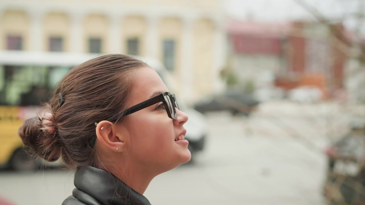 Primo piano di una giovane donna con capelli castani raccolti a chignon, che indossa occhiali neri e orecchini, in piedi all'aperto. La donna è vista di profilo, sorride leggermente mentre guarda in lontananza. Lo sfondo mostra