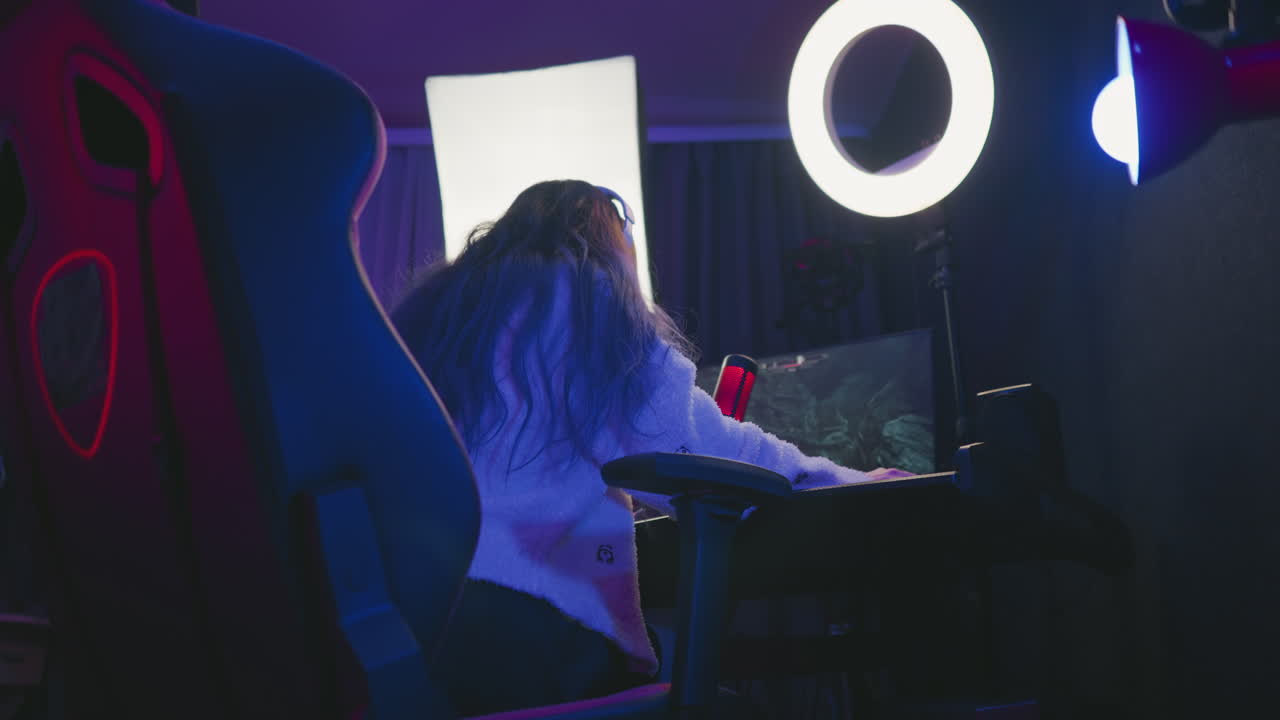 woman seated on gaming chair immersed in playing intense video game with large monitor and soft ambient lighting, professional setup including ring light and microphone visible in dim gaming room