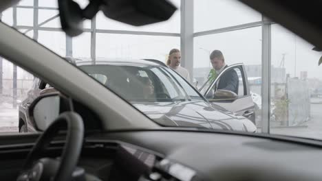 young car salesman showing to young couple new automobile at dealership salon.