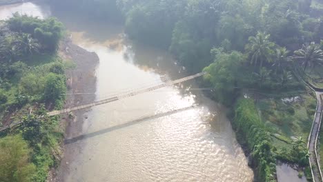 Vista-De-Drones-Del-Puente-Colgante-Sobre-El-Río-Con-Cruce-De-Motocicletas-En-él-Por-La-Mañana-Con-Rayos-De-Sol-Y-Niebla,-Java-Central,-Indonesia