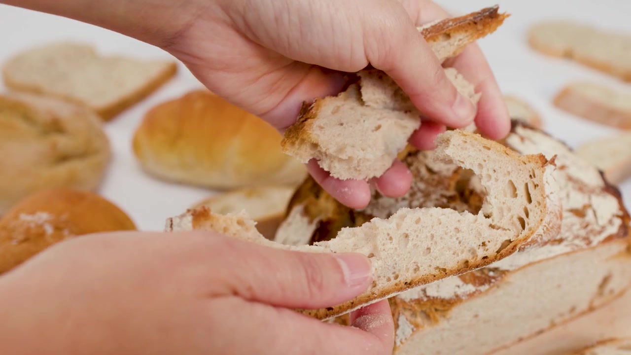 Close-up of hands tearing crusty sourdough bread on a white surface with various baked goods.