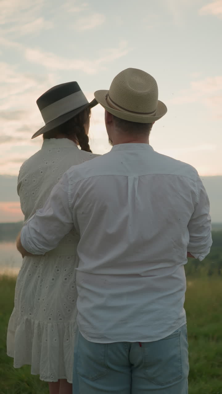 해가 지는 초원 언덕 위에 서서 하늘을 쳐다보는 연인들의 클로즈업. 색 셔츠, 모자, 청바지를 입은 남자가 여자를 허리에 부드럽게 붙잡고 있다. 그녀는 모자와 색 드레스를 입고 있다.