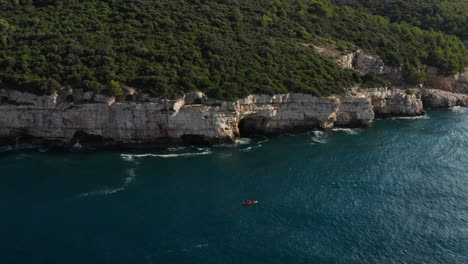 Luftaufnahme-Eines-Kajaks,-Das-Neben-Einer-Klippe-Und-Dichtem-Grün-In-Der-Nähe-Von-Pula-Istrien-In-Kroatien-Segelt