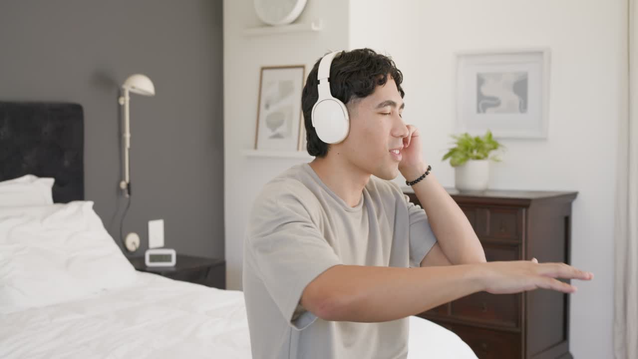 Asian African American man in 20s sitting on bed with white headset listening to music, singing