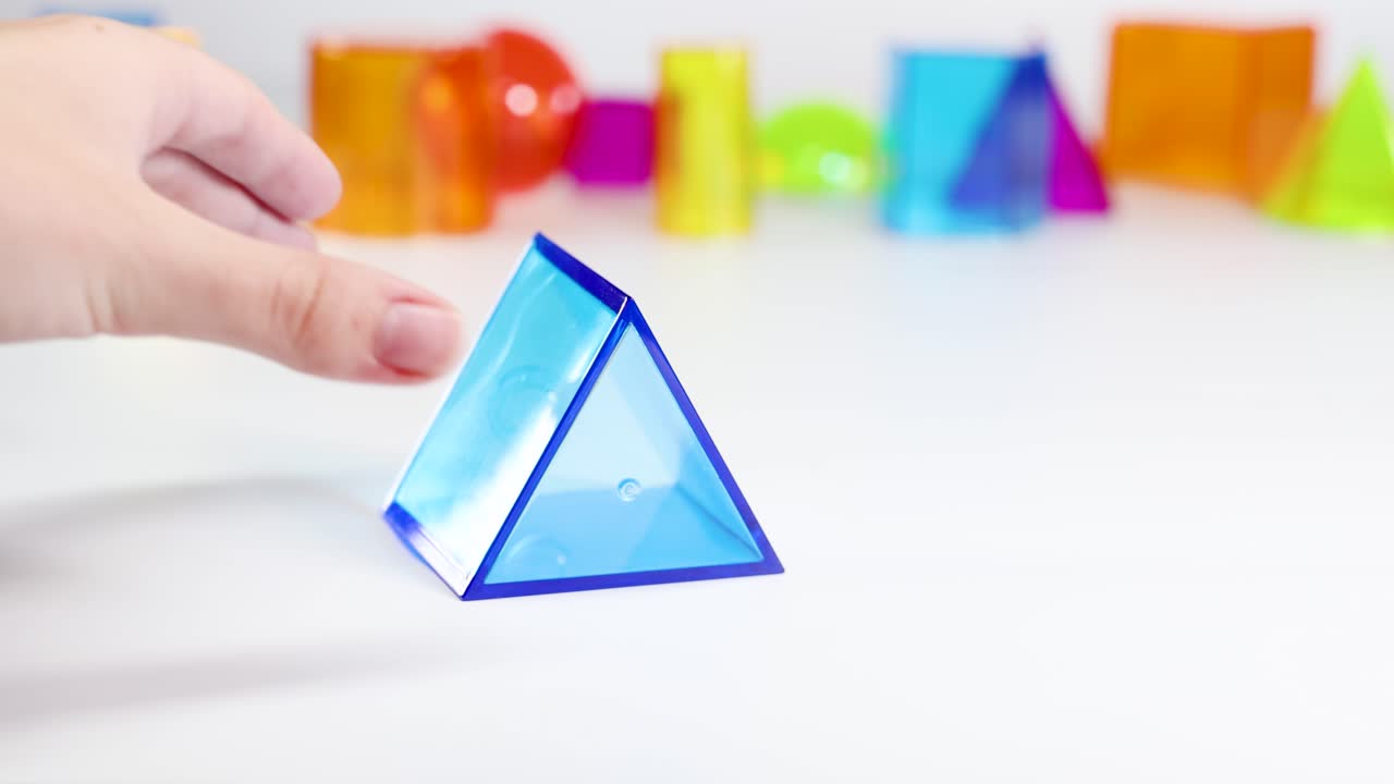 A hand manipulates a blue triangular prism among vibrant 3D shapes on a white surface, under bright lighting