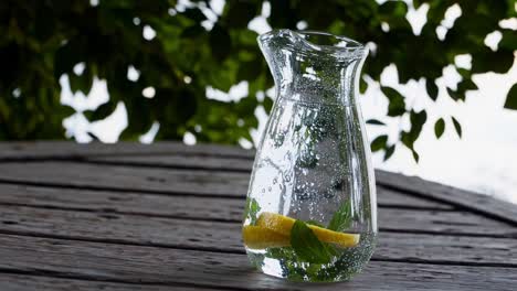 glass carafe with refreshing infused water, containing lemon slices and mint leaves, sits on a weathered wooden table, with a blurred backdrop of green foliage