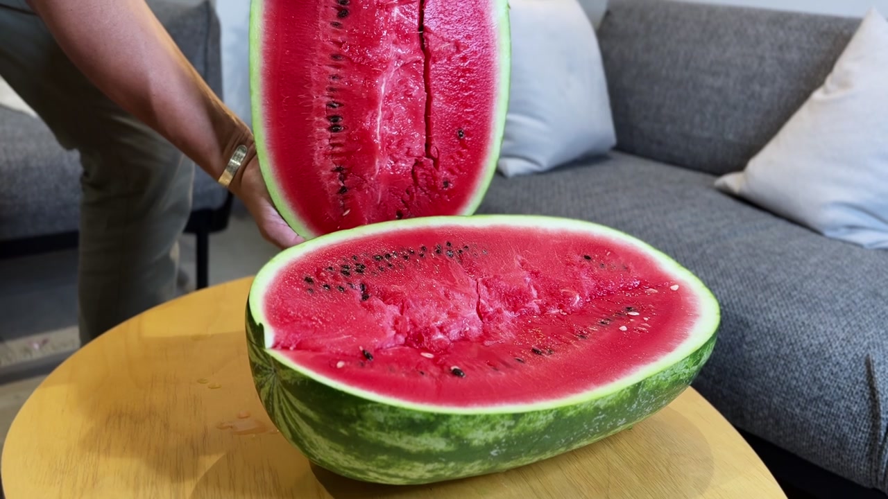 A person holds up half of a fresh, juicy red watermelon over a wooden table. The large cut fruit is displayed in a modern living room setting with a grey sofa in ambient daylight.